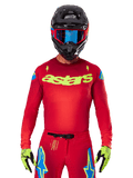 Alpinestars 2025 Supertech Maker Jersey in leuchtendem Rot und Gelb Fluo, mit gelbem „astars“-Branding auf der Brust und blauen Stern-Designs an den Schultern, getragen mit passender Hose, schwarzem Helm, blauer Brille und gelben Handschuhen, professionelle Motocross-Rennbekleidung