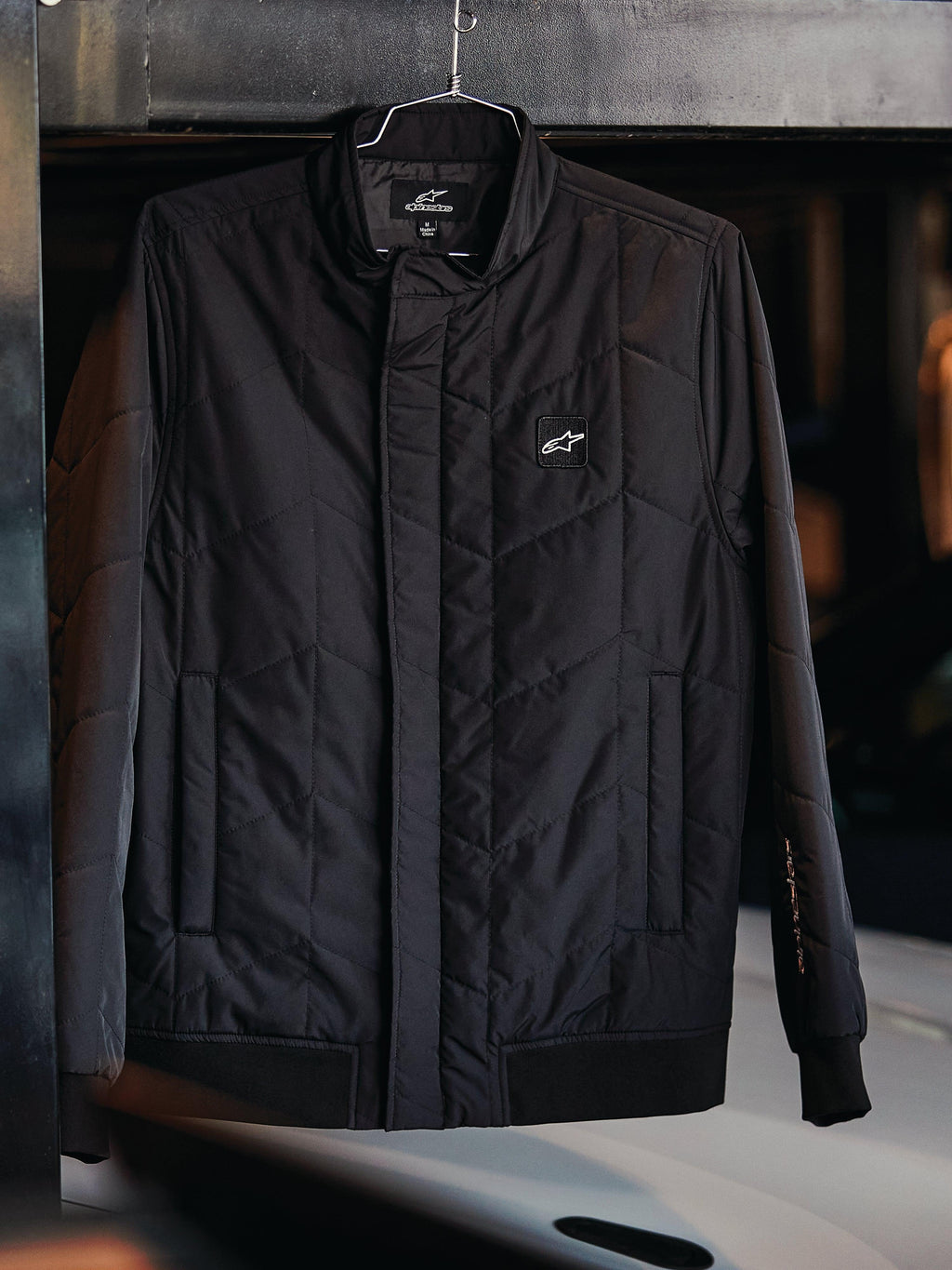 Alpinestars Precedent Jacke, gesteppte Sportswear-Jacke, Schwarz, mit schrägem Steppmuster, niedrigem Stehkragen mit verdecktem Reißverschluss, Logo-Patch auf der Brust und Rippbündchen an Ärmeln und Saum, Motorsport-inspiriertes Design