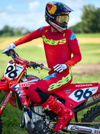 Motocross-Fahrer auf einem roten Honda-Motorrad im Alpinestars 2025 Supertech Maker Gear Set in Rot, bestehend aus einem roten Trikot mit neongrünem „astars“-Branding und blauen Schulterakzenten, passender roter Hose, roten Stiefeln, weißen Handschuhen und einem Red Bull-Helm, professionelle Rennausrüstung für Offroad-Performance