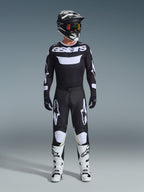 Motocross rider wearing complete racing gear, Alpinestars 2026 Racer Air Riway Jersey in Black and White with 'astars' branding on chest, matching black pants with white accents, white motocross boots, and black and white helmet with goggles, coordinated professional off-road racing outfit