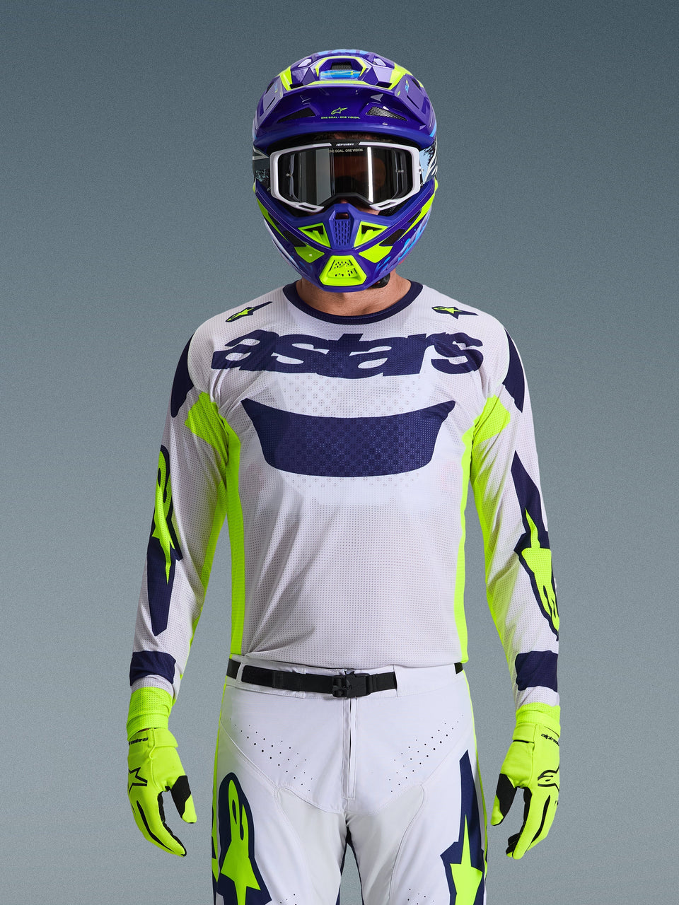 Alpinestars 2026 Racer Air Riway Trikot, Hellgrau mit dunkelblauem Astars-Logo, fluorgelbe und blaue Akzente an den Ärmeln, getragen von einem Motocross-Fahrer mit passendem lila und neongelbem Helm und Handschuhen, technisches Mesh-Gewebe für Offroad-Rennen.