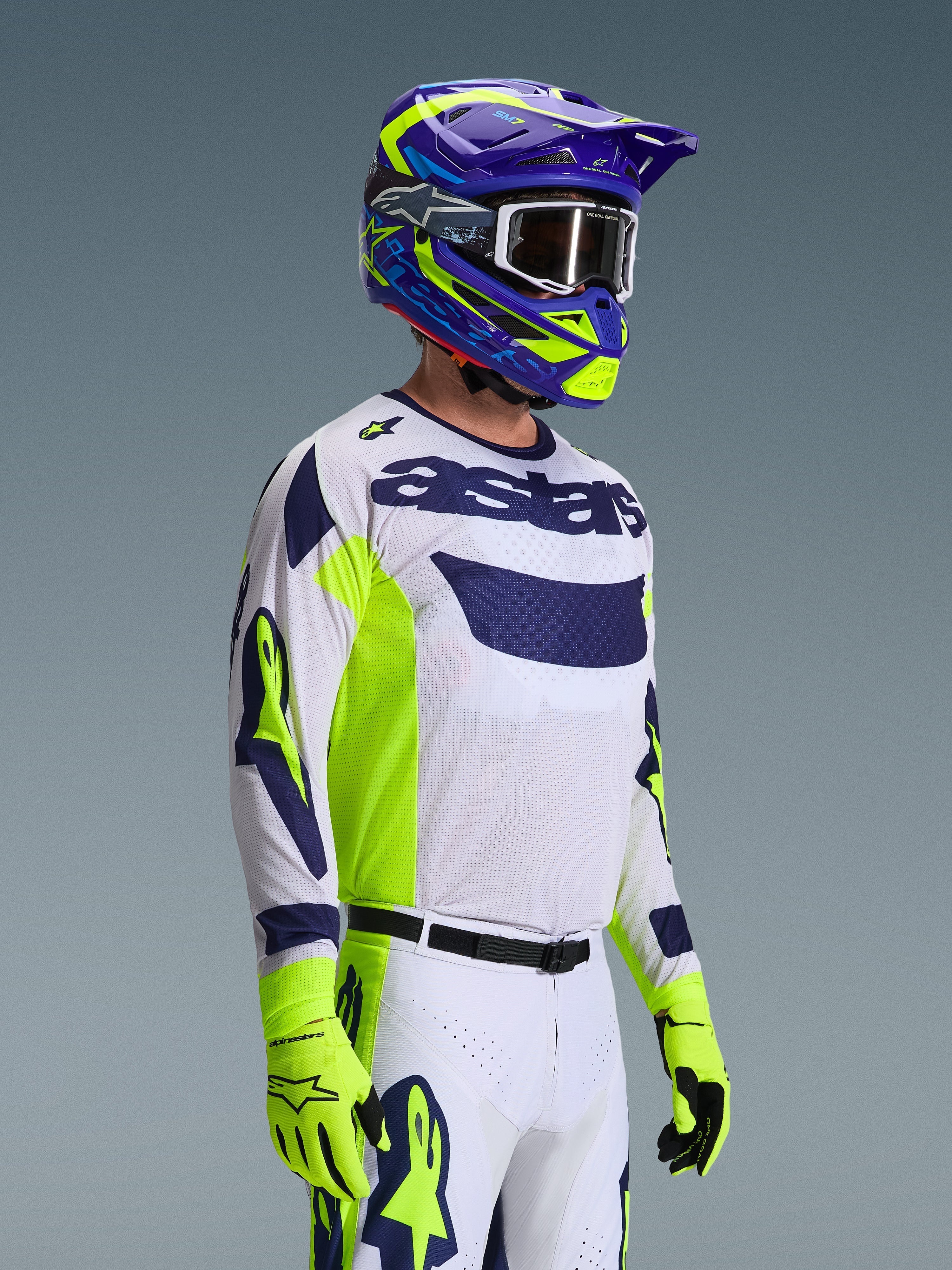 Motocross-Fahrer im Alpinestars 2026 Racer Air Riway Bekleidungsset, weißes Jersey mit marineblauem Logo und neongelben Seiteneinsätzen, weiße Hose mit passenden marineblauen und gelben Akzenten, lila und neongelber Helm mit weißer Brille, neongelbe Handschuhe, Hochleistungs-Offroad-Rennausrüstung