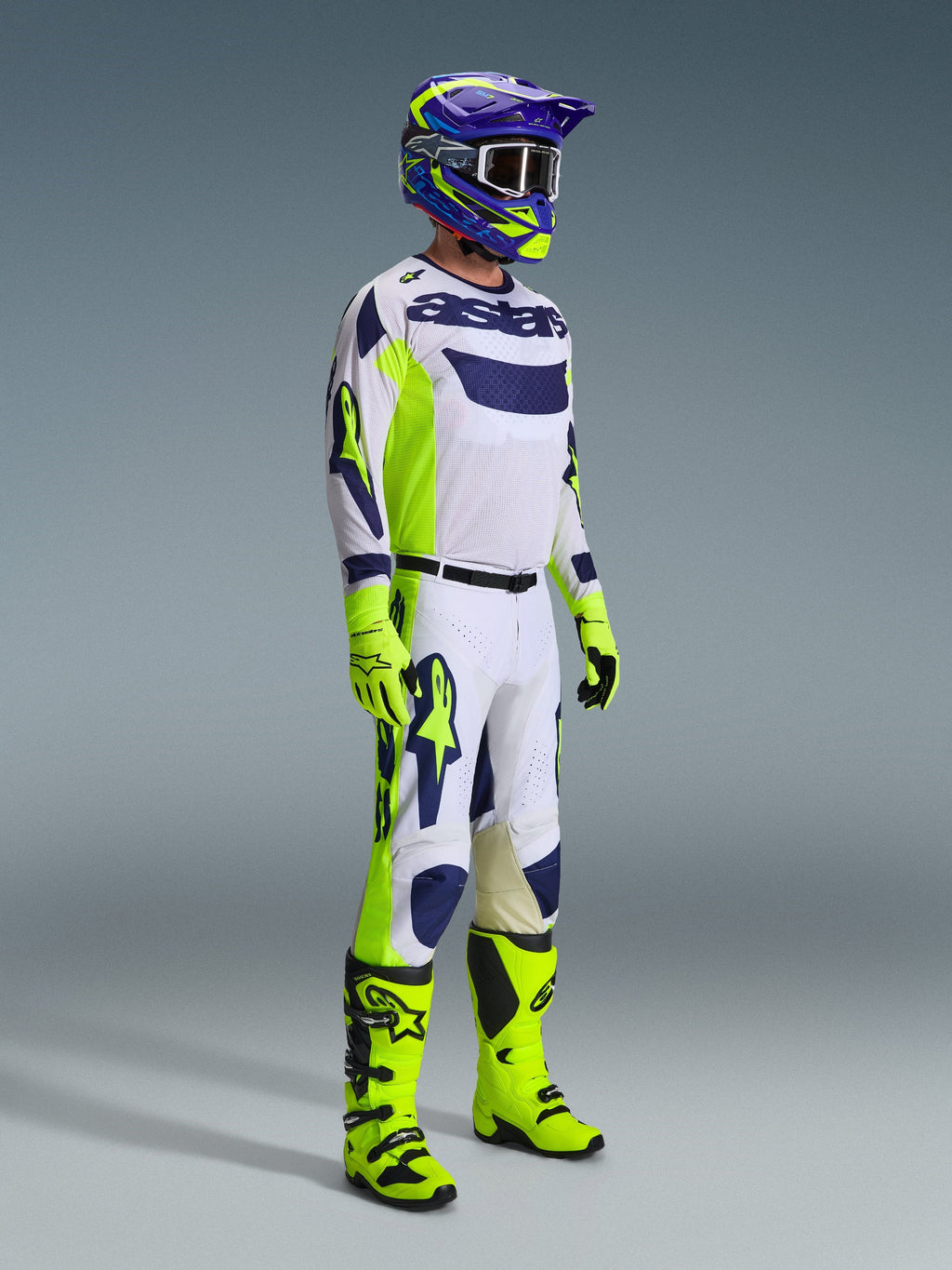 Motocross-Fahrer im Alpinestars 2026 Racer Air Riway Gear Set in Hellgrau, Neongelb und Blau. Das Set umfasst ein langärmeliges Mesh-Trikot, eine passende Rennhose, leuchtend neongelbe Handschuhen und Stiefel sowie einen lila-blauen Helm mit Schutzbrille, entwickelt für belüftete Offroad-Performance.
