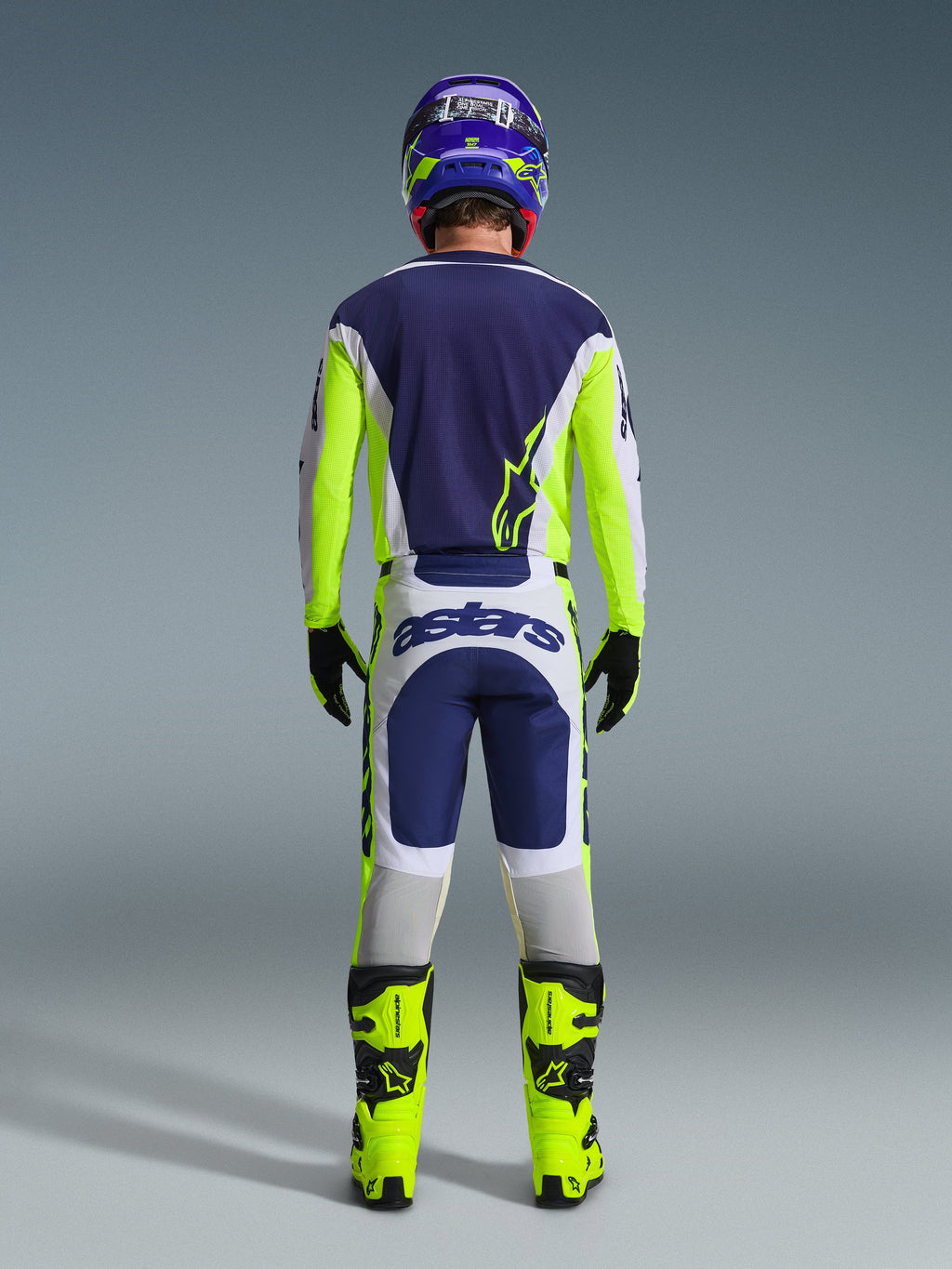 Motocross-Fahrer von hinten im Alpinestars 2026 Racer Air Riway Gear Set, blaues Trikot mit neongelben und hellgrauen Akzenten, weiß-blaue Hose mit neongelben Seiteneinsätzen, neongelbe Stiefel mit schwarzen Details, passender blau-gelber Helm, professionelle Ausrüstung für Offroad-Fahrten bei warmem Wetter
