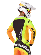 Motocross-Fahrer im Alpinestars 2025 Techstar Melt Jersey, Rückansicht, Neongelb mit orangen Ärmeln und einem großen schwarzen Grafikpanel, passende Motocross-Hose mit markantem Branding, weißer Alpinestars-Helm und orange Brille, professionelle Offroad-Rennausrüstung