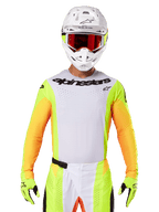 Alpinestars 2025 Techstar Melt Trikot, Motocross-Rennbekleidung, weiße Brust mit neongelben und orangen Ärmeln, Fahrer mit weißem Helm und roter Brille, neongelbe Handschuhe mit schwarzen Logos, lebendige Farbkombination für Offroad-Performance