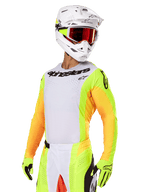 Motocross-Fahrer im Alpinestars 2025 Techstar Melt Gear Set, weißes Trikot mit leuchtend gelben und orangefarbenen Ärmeln, schwarzes 'Alpinestars'-Branding auf der Brust, passende neongelbe Handschuhe, weißer Helm mit rot getönter Brille, professionelle Offroad-Rennausrüstung