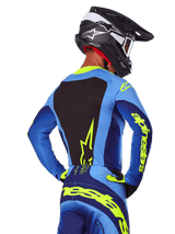 Rückansicht eines Motocross-Fahrers im Alpinestars 2025 Techstar Melt Jersey in Blau und Gelb Fluo, mit schwarzem Rückeneinsatz und markantem gelbem Branding auf den Ärmeln, kombiniert mit passender Motocross-Hose und einem schwarzen Carbon-Helm, Performance-Offroad-Rennausrüstung