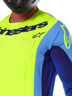 Alpinestars 2025 Techstar Melt Jersey, Motocross Jersey, Yellow Fluo and Blue, featuring laser-cut perforations and bold logo on the chest, athletic fit for off-road racing, close-up view with a black helmet