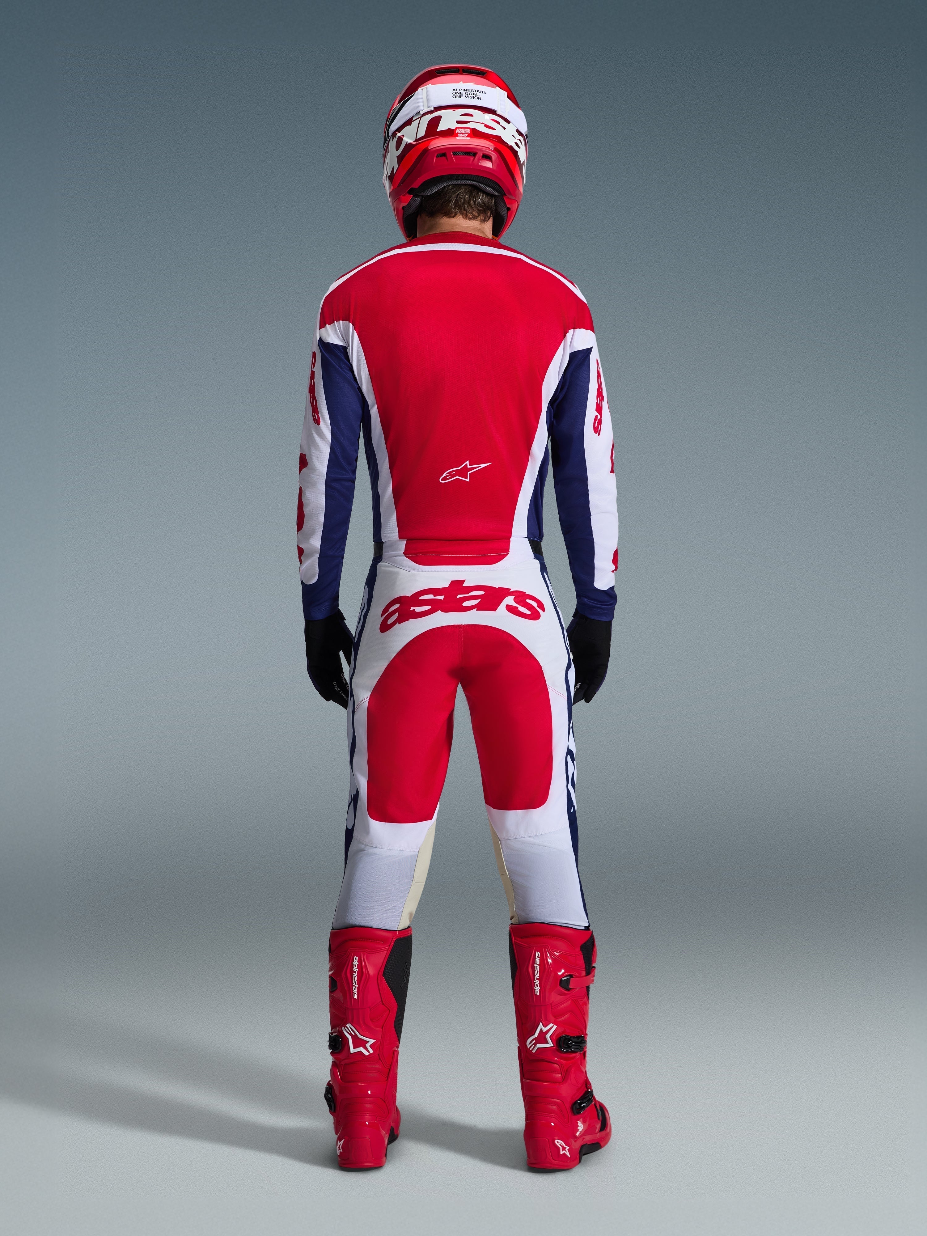 Motocross-Fahrer in kompletter Rennausrüstung von hinten, Alpinestars 2026 Racer Riway Jersey in Weiß-Blau-Rot, passende weiß-rote Hose mit markantem 'astars'-Branding, rote Stiefel und rot-weißer Helm, abgestimmtes Offroad-Rennoutfit.