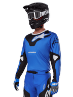 Alpinestars 2025 Racer Veil Ausrüstungsset, blaues Motocross-Trikot mit schwarz-weißen Grafiken, getragen mit passender Hose, schwarzem Helm, weißer Brille und blauen Handschuhen, technische Bekleidung für den Offroad-Rennsport