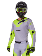 Motocross-Fahrer im Alpinestars 2025 Racer Veil Ausrüstungsset in Grau und Neongrün, hellgraues Trikot und Hose mit markanten neongelb-grünen Einsätzen und schwarzen Alpinestars-Logos, kombiniert mit passenden Handschuhen und einem schwarzen Helm mit neongrüner Brille, professionelle Offroad-Rennbekleidung.