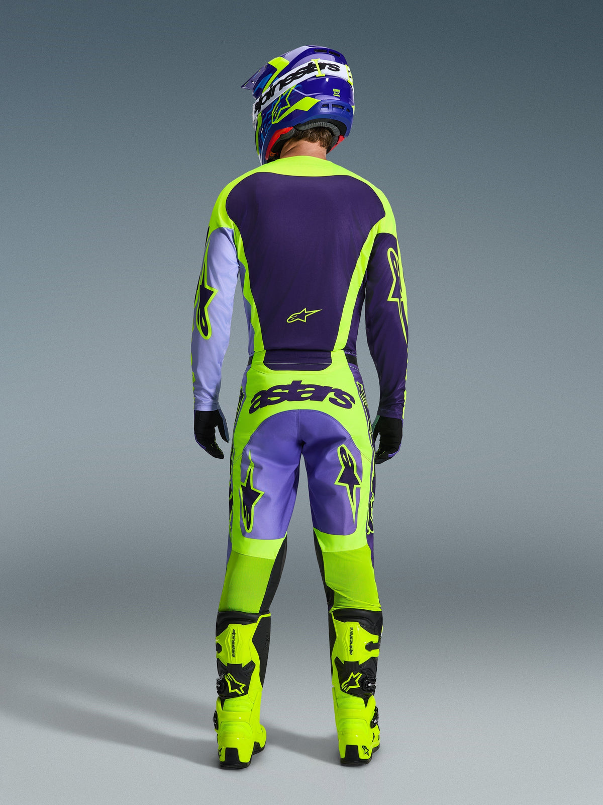 Motocross rider from behind wearing the Alpinestars 2026 Racer Portl Jersey in Purple and Yellow Fluo, paired with matching motocross pants, fluo yellow boots, and a blue and yellow helmet, showcasing a complete off-road gearset.