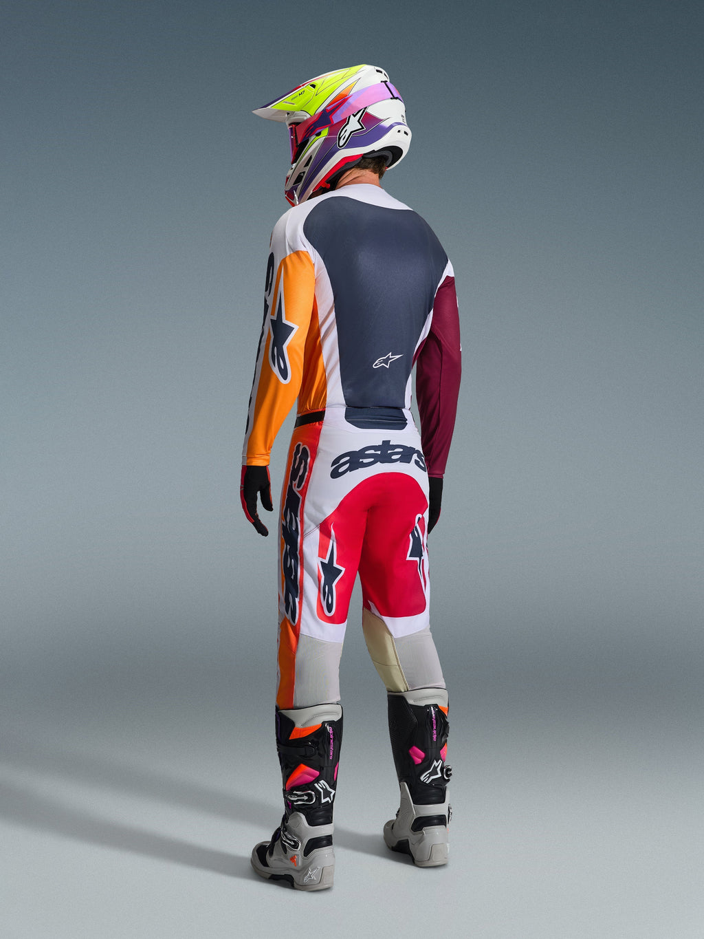 Motocross rider from behind wearing the Alpinestars 2026 Racer Portl Gear Set, featuring a color-blocked jersey with orange and maroon sleeves, white pants with red panels and bold 'astars' branding, multi-colored helmet, and gray boots, professional off-road racing outfit.