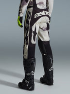 Alpinestars 2026 Racer Lahnd Hose, Motocross-Hose, Sand-Camo und Grau, Rückansicht mit markantem weißen Branding an Gesäß und Beinen, kombiniert mit schwarzen Motocross-Stiefeln und gelben Handschuhen, professionelle Offroad-Rennausrüstung