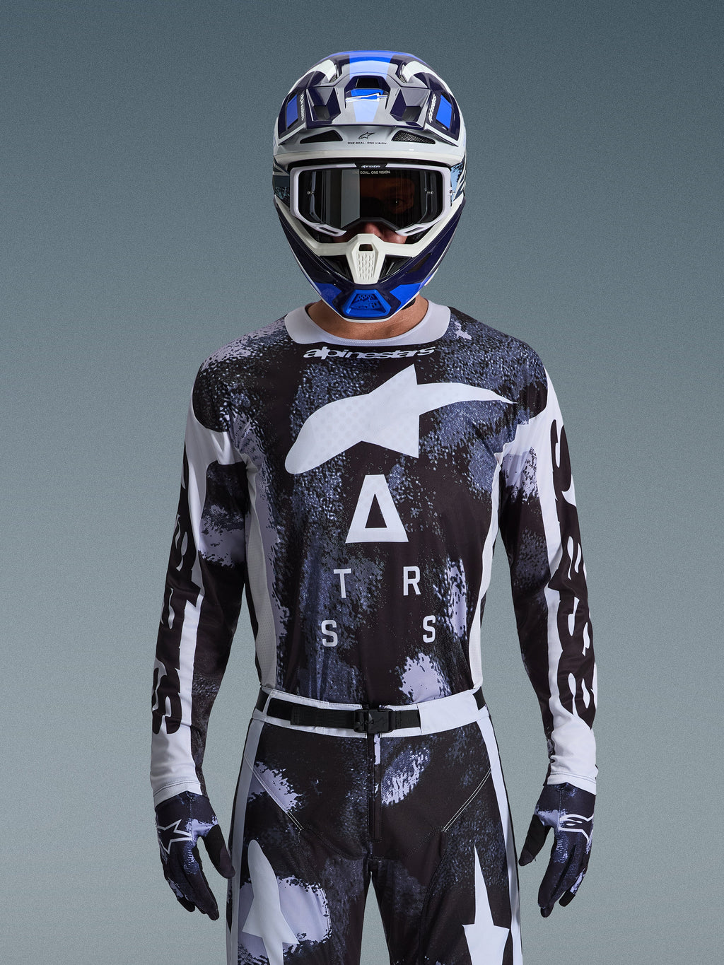 Motocross-Fahrer im Alpinestars 2026 Racer Lahnd Bekleidungsset, schwarz-weißes Jersey mit abstraktem Camouflage-Muster, großen weißen Logos und „ATRS“-Schriftzug, passende Hose und Handschuhe, blau-weißer Helm mit Schutzbrille, Hochleistungs-Rennausrüstung für Offroad-Motocross
