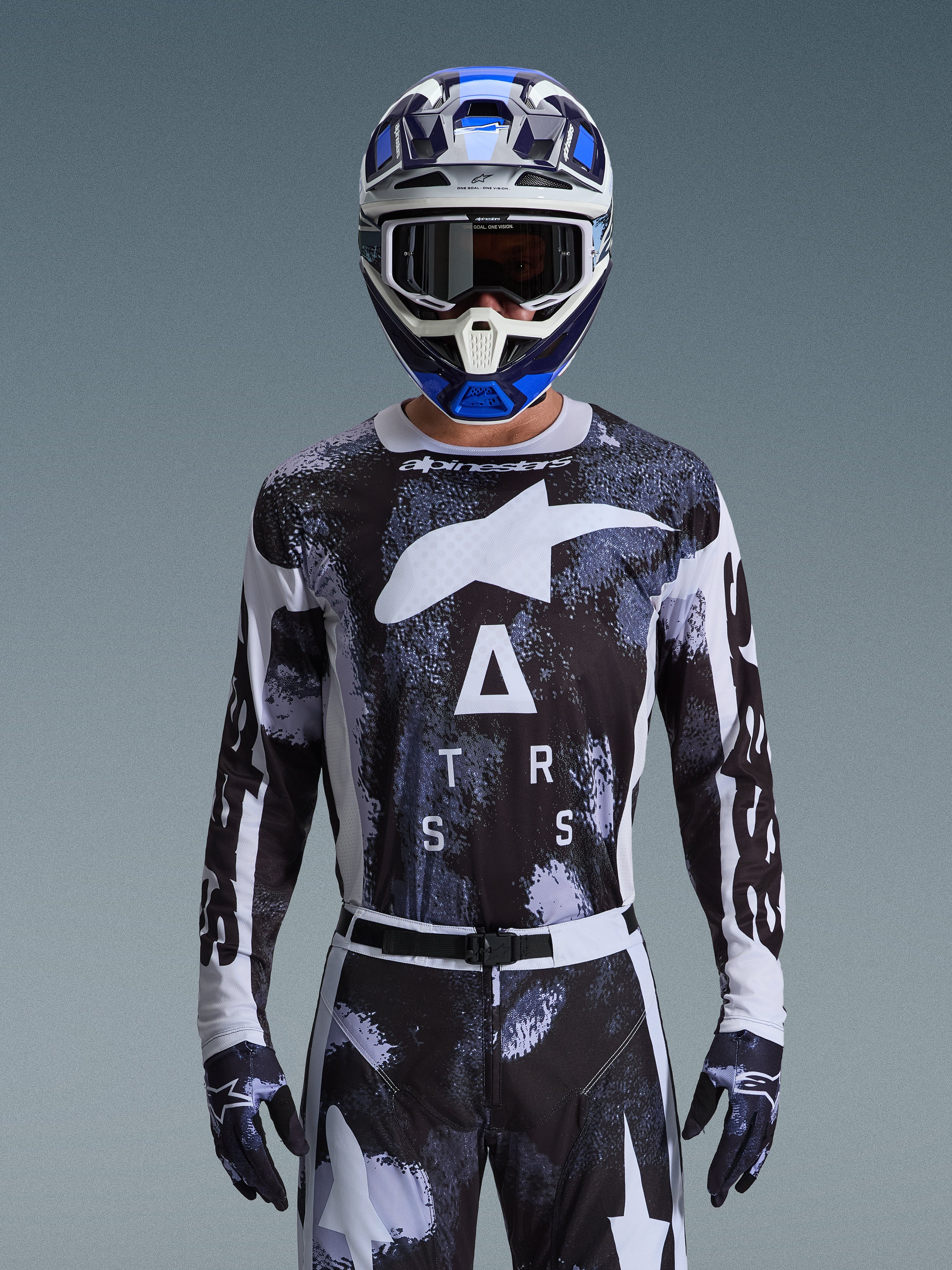 Motocross-Fahrer im Alpinestars 2026 Racer Lahnd Bekleidungsset, schwarz-weißes Jersey mit abstraktem Camouflage-Muster, großen weißen Logos und „ATRS“-Schriftzug, passende Hose und Handschuhe, blau-weißer Helm mit Schutzbrille, Hochleistungs-Rennausrüstung für Offroad-Motocross