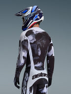 Alpinestars 2026 Racer Lahnd Gear Set, Motocross-Fahrer von hinten in Trikot und Hose mit schwarz-weißer, gesprenkelter Grafik, kombiniert mit einem blau-weißen Alpinestars-Helm und schwarzen Handschuhen, Performance-Offroad-Rennausrüstung