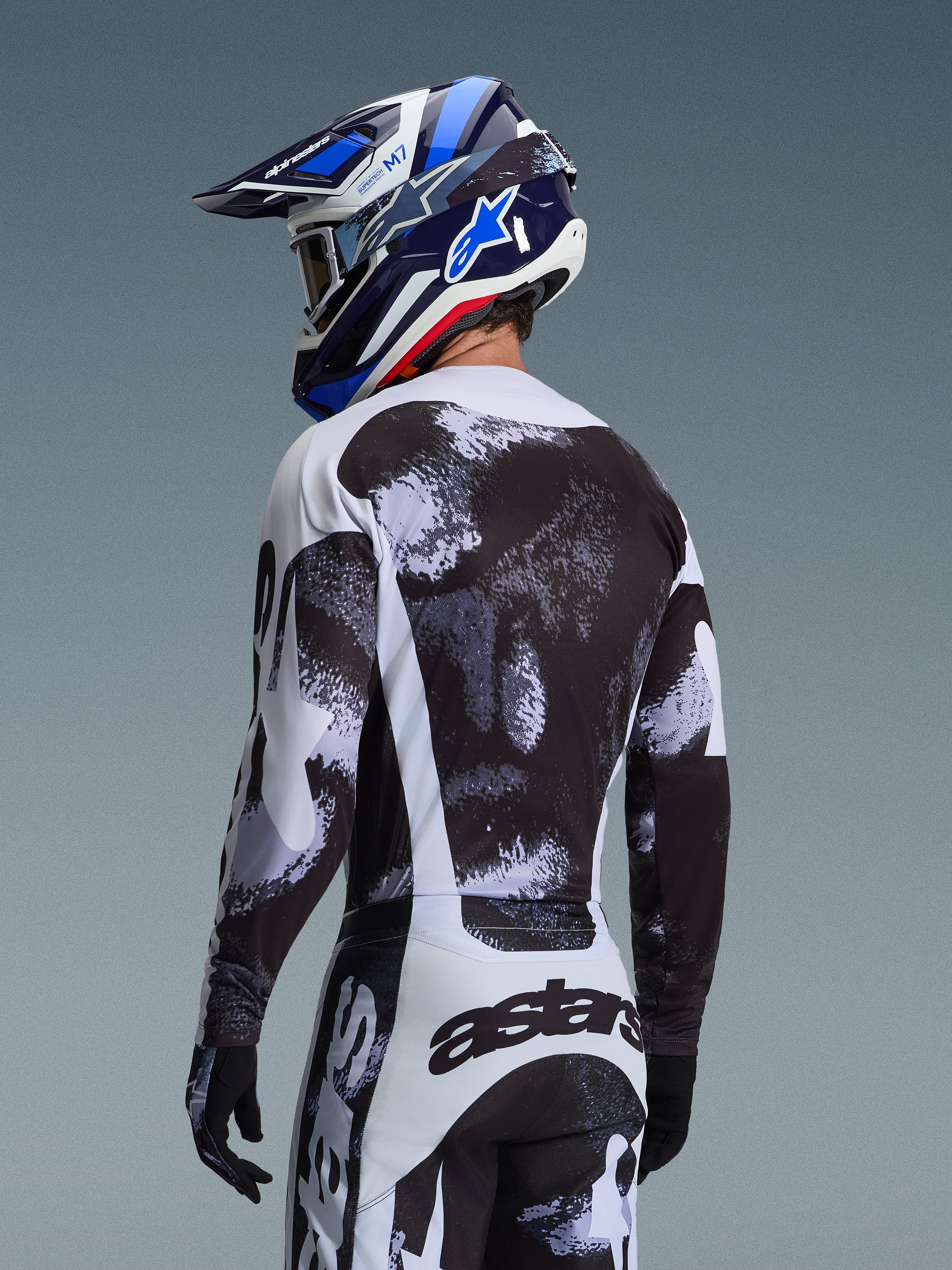 Alpinestars 2026 Racer Lahnd Gear Set, Motocross-Fahrer von hinten in Trikot und Hose mit schwarz-weißer, gesprenkelter Grafik, kombiniert mit einem blau-weißen Alpinestars-Helm und schwarzen Handschuhen, Performance-Offroad-Rennausrüstung