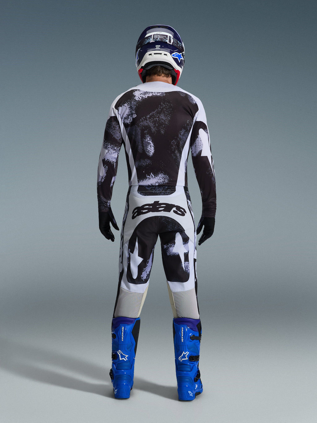 Motocross-Fahrer in der Rückansicht im Alpinestars 2026 Racer Lahnd Gear Set, schwarz-weiß abstrakt gemustertes Jersey und Hose, „astars“-Branding auf dem Gesäß, kombiniert mit blauen Stiefeln und passendem Helm, professionelle Offroad-Rennbekleidung.