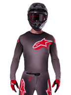 Alpinestars 2025 Supertech Lipan Trikot, Motocross-Jersey, Smoke und Rot, mit einem großen rot-weißen Brustlogo, getragen mit einem schwarzen Helm und rot-schwarzen Handschuhen, athletische Rennpassform mit lasergeschnittenen Perforationen