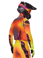 Motocross rider seen from a rear-side view wearing the Alpinestars 2025 Supertech Lipan Gear Set, featuring a yellow, orange, and red gradient with black and pink branding, black helmet with pink accents and orange goggles, professional off-road racing attire.