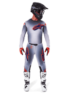 Motocross rider wearing the 2025 Supertech Lipan Gear Set in Gray Orange Fluo, featuring a gray jersey with orange accents and a navy Alpinestars logo, matching gray pants with large thigh logos, a matte black helmet, white and black boots, and red and black gloves, complete pro-level racing outfit.