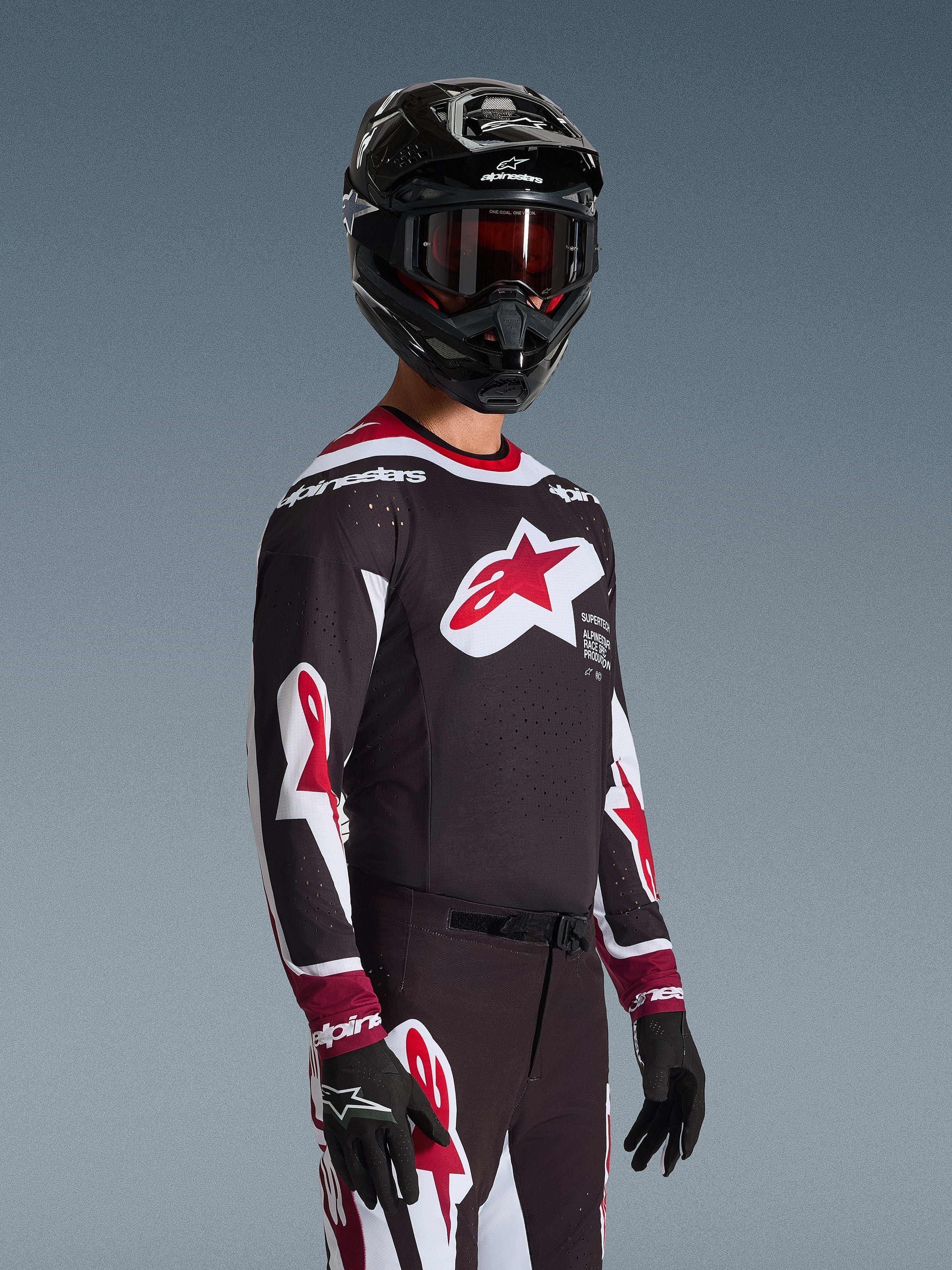 Alpinestars 2026 Supertech AFD Vista Trikot, Motocross-Fahrer in kompletter Rennausrüstung, schwarzes Trikot mit roten und weißen Akzenten, großes Logo auf der Brust, lasergeschnittene Perforationen, kombiniert mit passender Hose, schwarzem Helm und roter Brille, abgestimmtes Outfit für den Offroad-Rennsport
