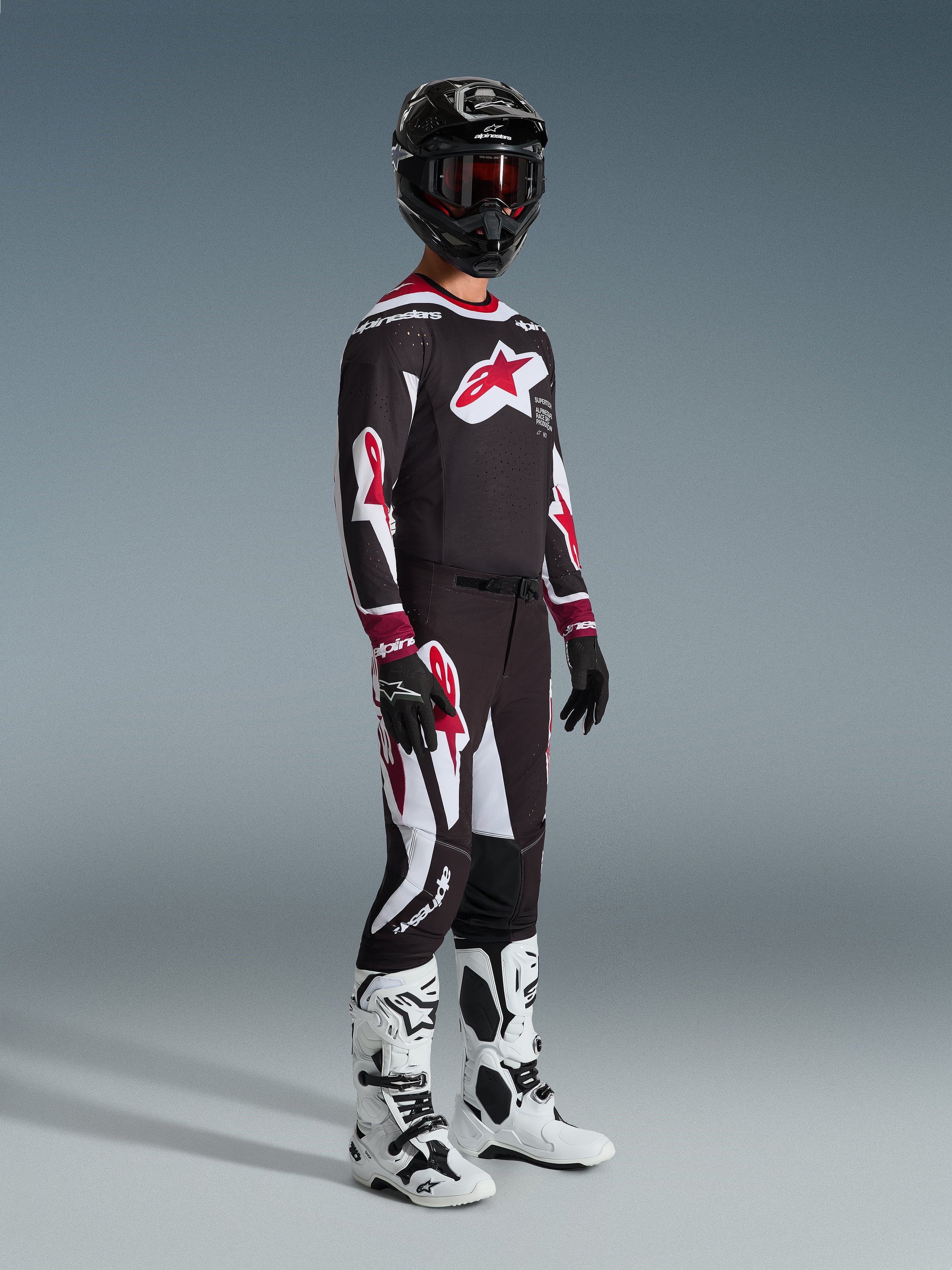 Alpinestars 2026 Supertech AFD Vista Bekleidungsset, Motocross-Fahrer in kompletter Rennausrüstung, schwarzes Jersey mit großem roten Stern-Logo auf der Brust, passende schwarze Hose mit roten und weißen Details, weiß-schwarze Motocross-Stiefel, schwarzer Helm mit roter Brille, professionelles Motocross-Rennoutfit
