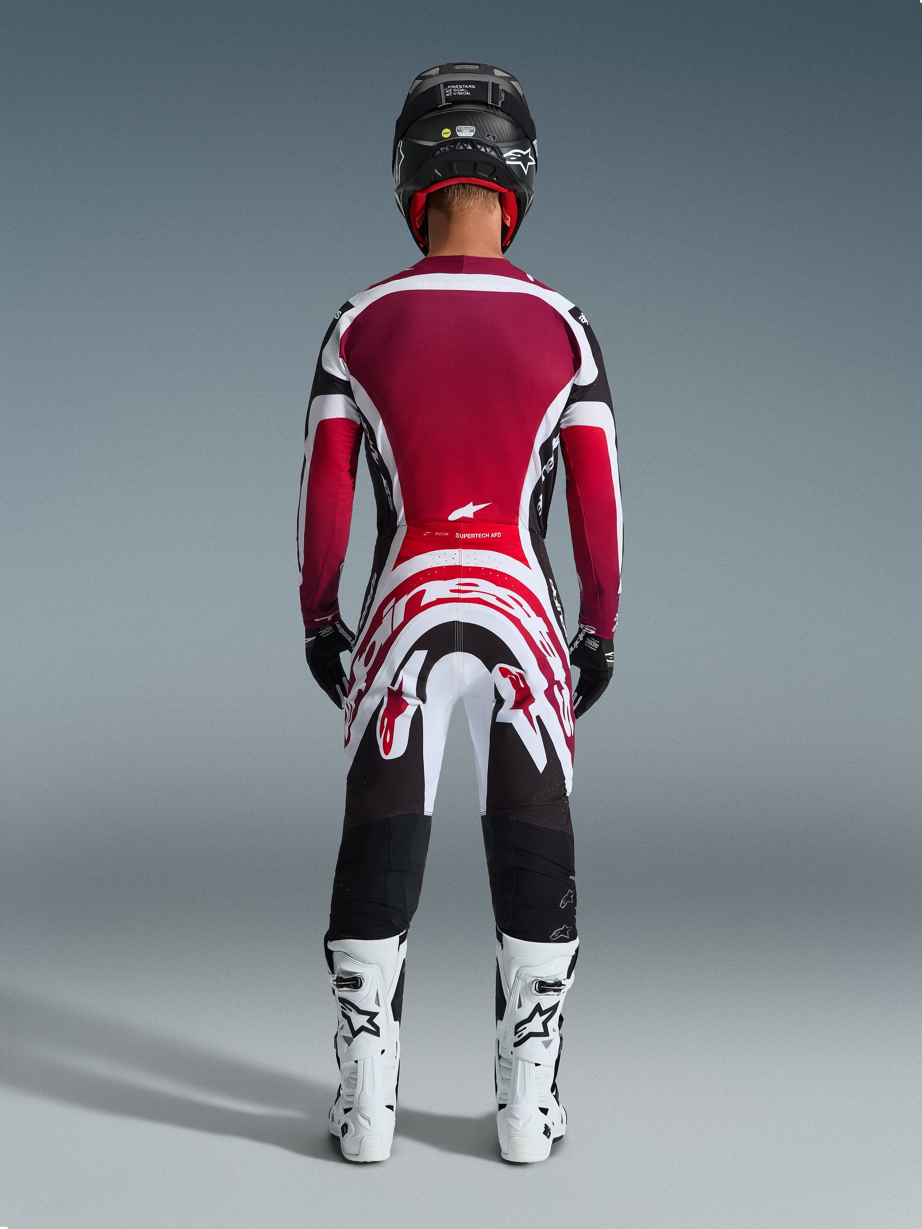 Alpinestars 2026 Supertech AFD Vista Gear Set, Rückansicht eines Motocross-Fahrers in professioneller Rennausrüstung, burgunderrotes und rotes Jersey mit weißen und schwarzen Akzenten, passende Hose mit auffälligen Grafiken, weiße Stiefel und schwarzer Helm, abgestimmtes Motocross-Rennoutfit.