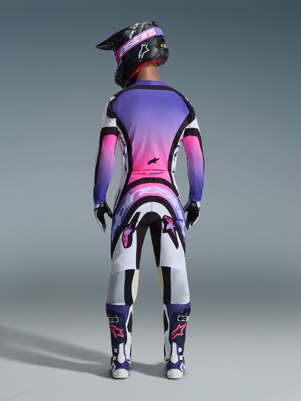 Motocross-Fahrer in der Rückansicht mit dem 2026 Supertech AFD Vista Trikot, Weiß, Lila und Schwarz mit pinkfarbenen Farbverlauf-Akzenten, passende Motocross-Hose, schwarzer Helm mit pinker Brille und weiß-lila Stiefel, komplettes professionelles Rennausrüstungs-Set