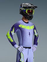 Motocross rider wearing the 2026 Alpinestars Supertech Scenz Jersey, Purple and Yellow Fluo, matching purple pants with fluo accents, black helmet with yellow goggles, and purple racing gloves, coordinated professional gear for off-road racing