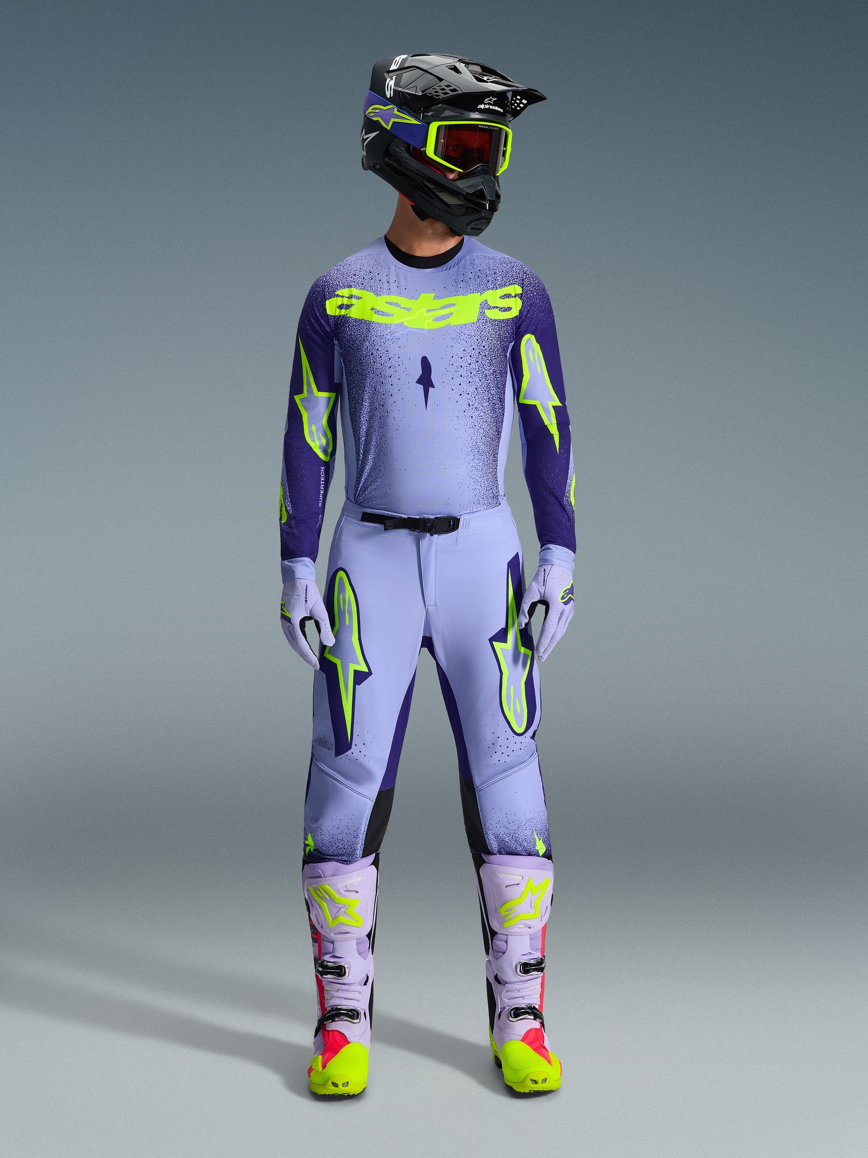 Motocross-Fahrer in kompletter Alpinestars 2026 Supertech Scenz Rennausrüstung, Trikot in Lila und Gelb Fluo mit passender Hose, schwarzer Helm mit gelber Schutzbrille, helllila und neonfarbene Stiefel, professionelles Outfit mit athletischer Passform für den Offroad-Rennsport