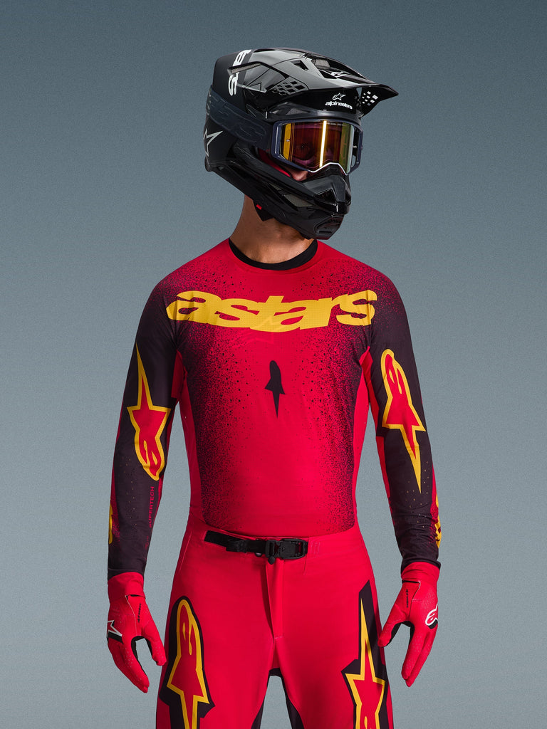 Alpinestars 2026 Supertech Scenz Trikot, Motocross-Trikot, Rot, Schwarz und Gelb, getragen von einem Fahrer mit einem passenden schwarzen Helm und roten Handschuhen, mit einem markanten gelben 'astars'-Logo auf der Brust und einem schwarzen, gesprenkelten Farbverlaufsmuster über den Schultern.