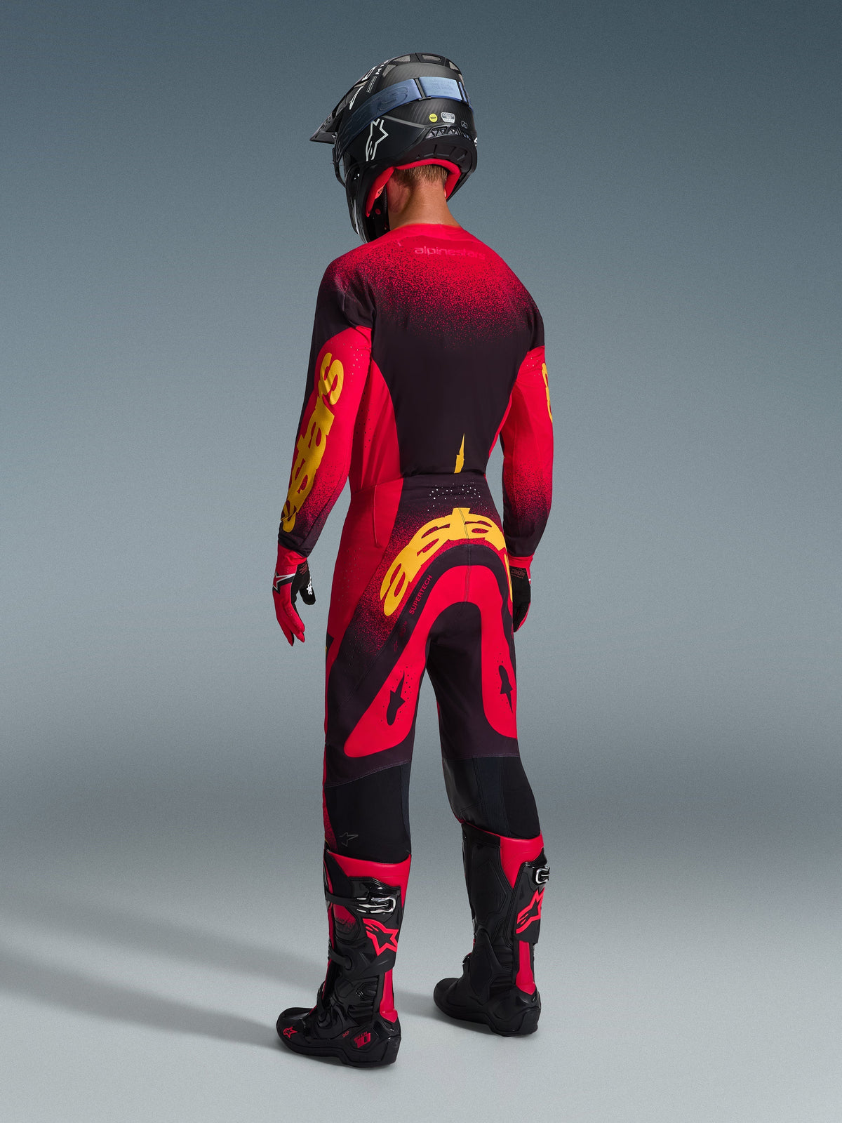 Motocross rider from the rear wearing complete racing gear, Red jersey with black and yellow accents, matching red and black pants with a large yellow Alpinestars logo, black helmet, black and red boots, red and black gloves, coordinated outfit for professional off-road racing
