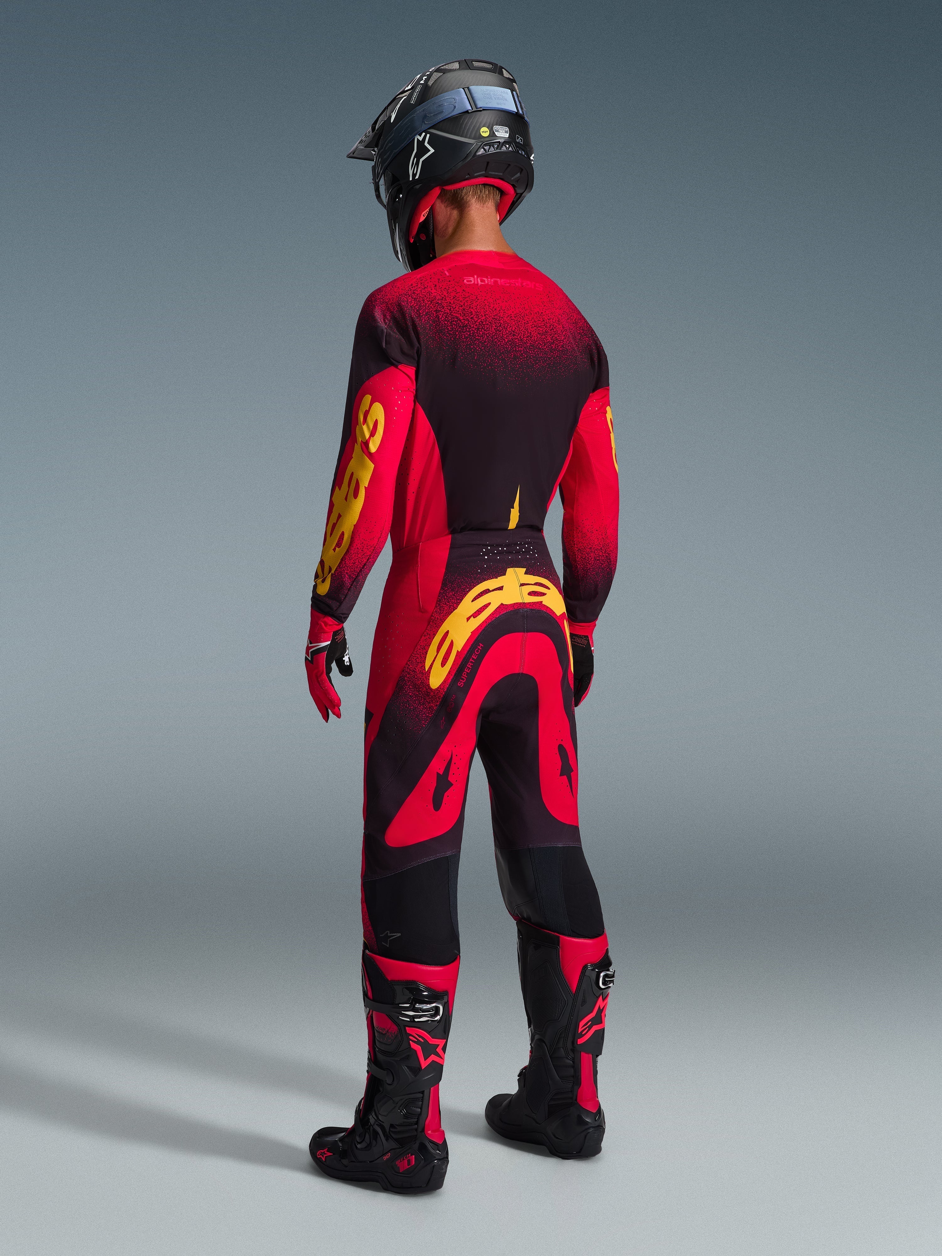 Motocross rider from the rear wearing complete racing gear, Red jersey with black and yellow accents, matching red and black pants with a large yellow Alpinestars logo, black helmet, black and red boots, red and black gloves, coordinated outfit for professional off-road racing