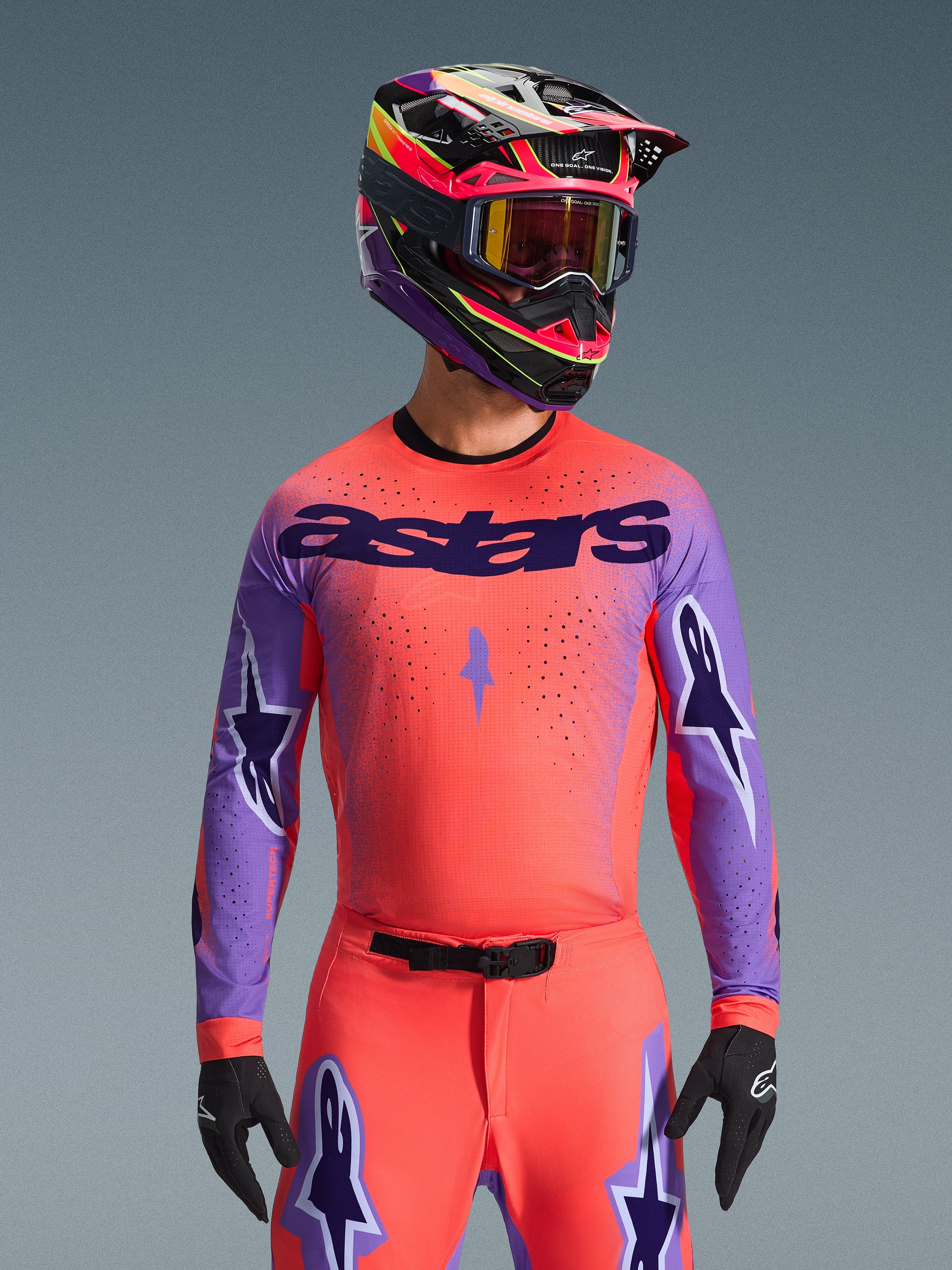 Motocross rider wearing the Alpinestars 2026 Supertech Scenz Gear Set, Vibrant pinkish-orange jersey with purple sleeves and 'astars' branding, matching pants with purple star accents, multi-colored racing helmet with goggles, black gloves, professional off-road racing gear