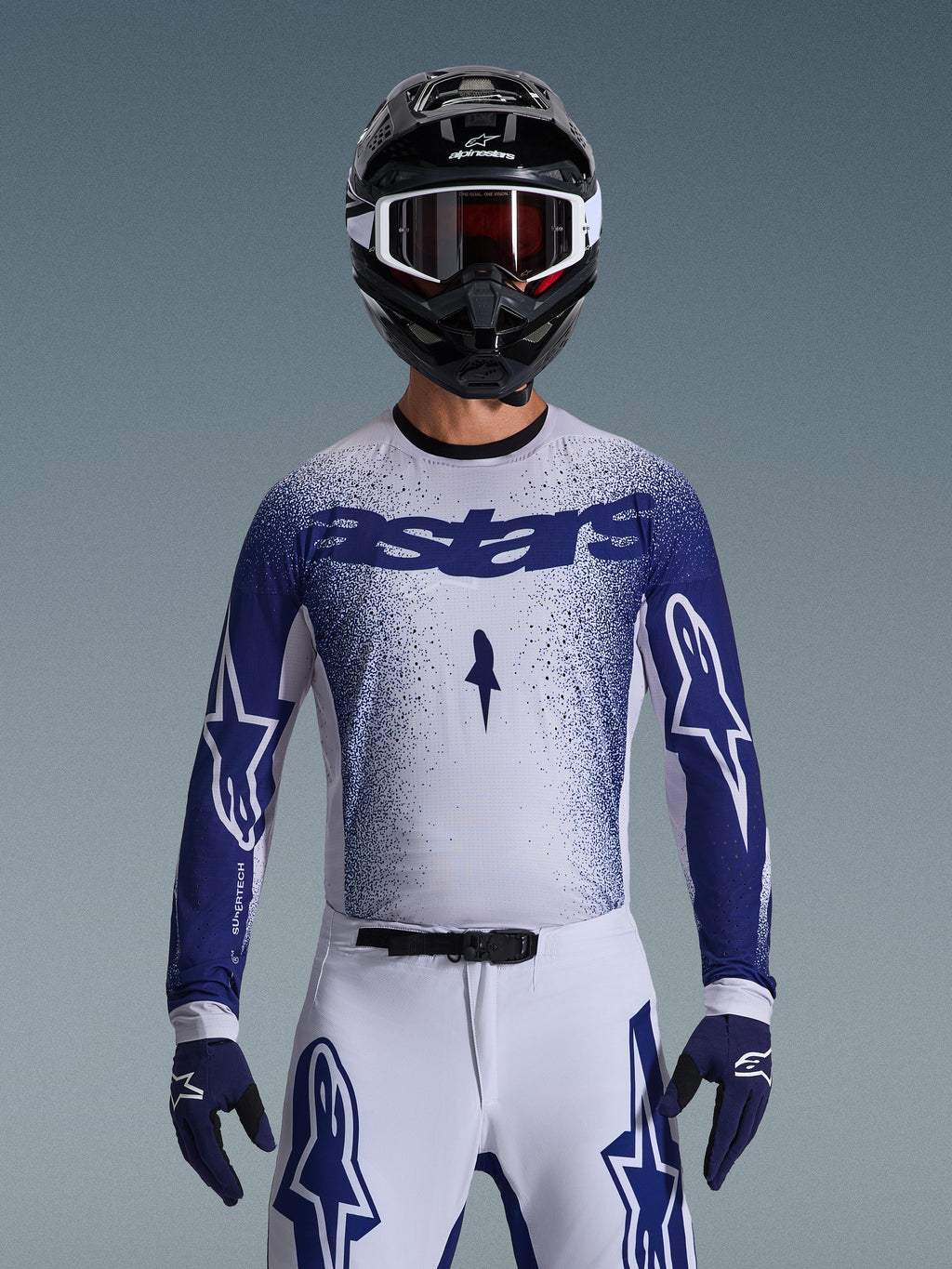 Motocross-Fahrer im Alpinestars 2026 Supertech Scenz Trikot, Hellgrau und Dunkelblau mit gesprenkeltem Farbverlauf und markantem „astars“-Branding. Getragen mit passender weißer Motocross-Hose, dunkelblauen Handschuhen und schwarzem Helm mit Schutzbrille, koordiniertes Outfit für professionellen Offroad-Rennsport.