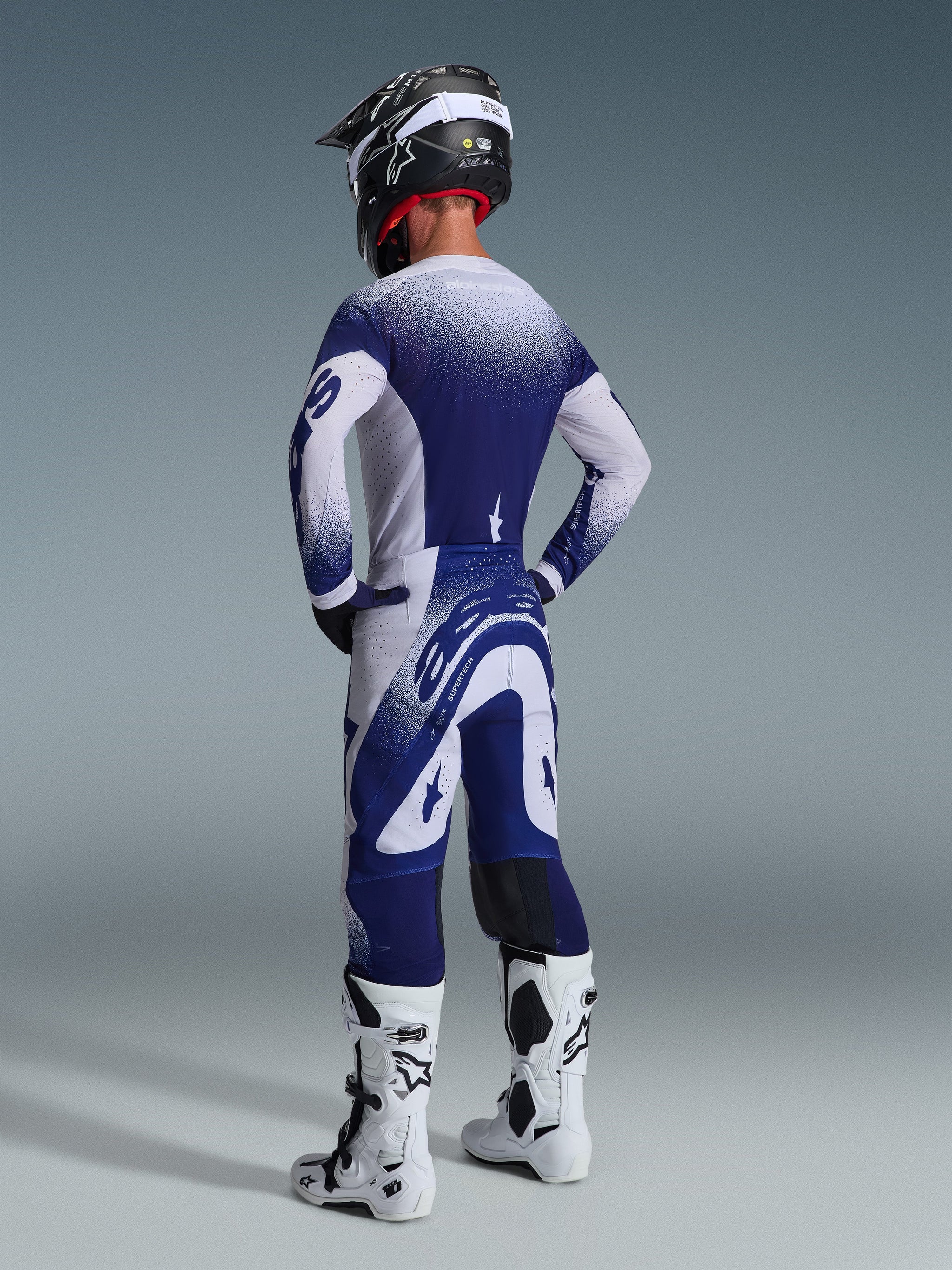 Rückansicht eines Motocross-Fahrers im Alpinestars 2026 Supertech Scenz Trikot in Hellgrau und Dunkelblau mit gesprenkeltem Farbverlaufsdesign, kombiniert mit passender Hose und weißen Motocross-Stiefeln, professionelle athletische Rennausrüstung.
