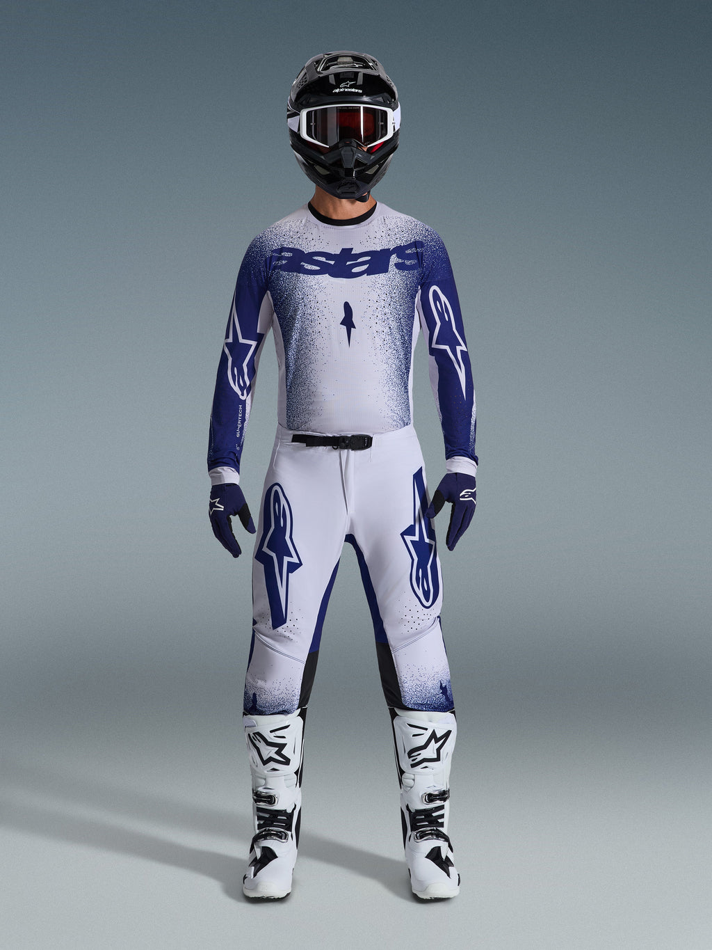 Motocross-Fahrer in kompletter Rennausrüstung, 2026 Supertech Scenz Trikot in Hellgrau und Dunkelblau, passende hellgraue Hose mit blauen Logos, schwarz-weißer Helm mit roter Brille, weiß-schwarze Stiefel, dunkelblaue Handschuhe, abgestimmtes Outfit für den Offroad-Rennsport
