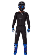 Alpinestars 2025 Racer Graphite Gear Set, Motocross rider wearing complete racing gear, black long-sleeve jersey with gray Alpinestars logos, black pants, black helmet with blue goggles, blue and gray gloves, blue and black boots, coordinated outfit for off-road motocross racing