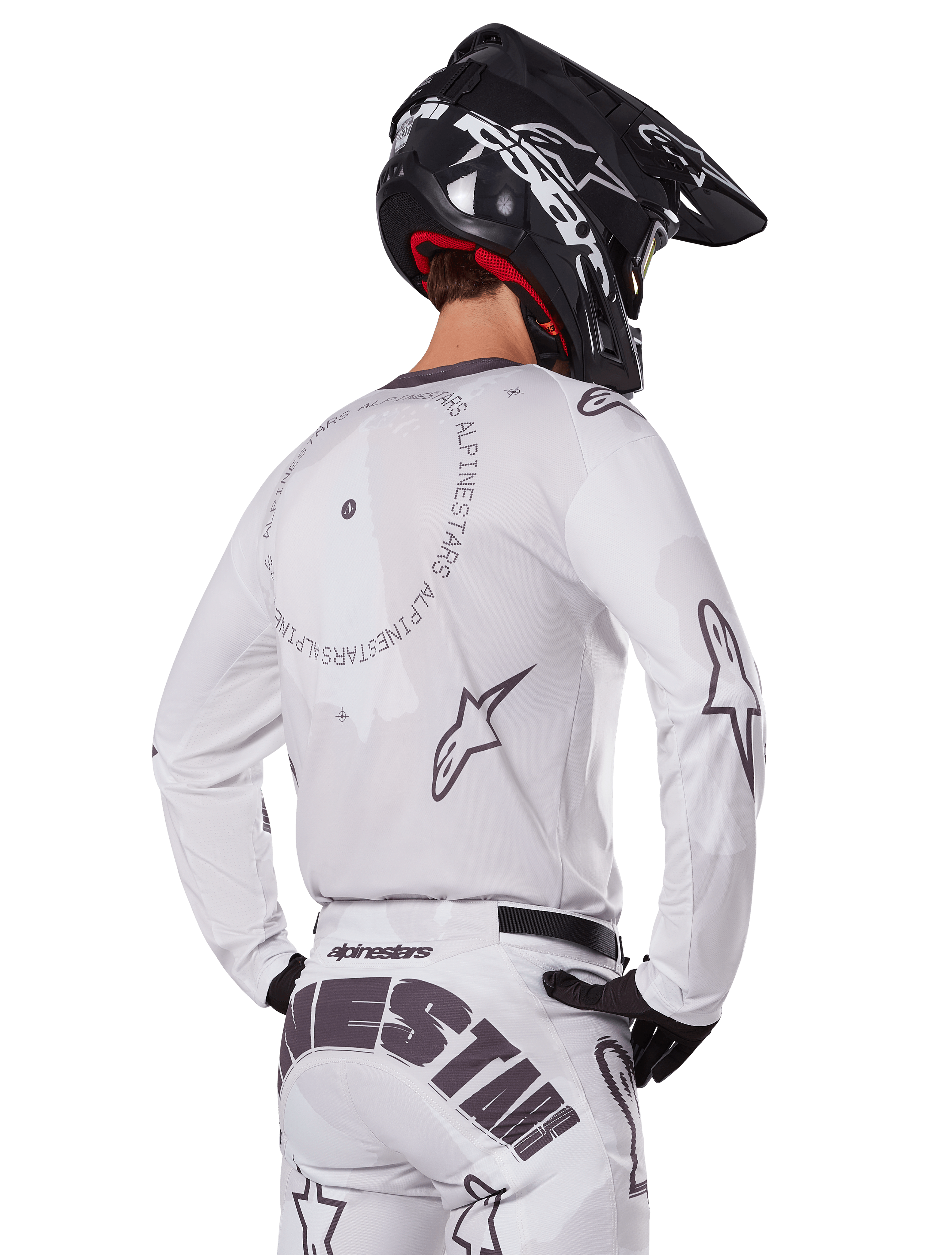 Motocross-Fahrer in der Rückansicht trägt das Alpinestars 2025 Racer Hollow Gear Set in Weiß, weißes Langarmtrikot mit grauen Sternlogos und kreisförmigen Grafiken, passende weiße Hose mit markantem Branding am Gesäß, schwarzer Helm, abgestimmte Offroad-Rennausrüstung.