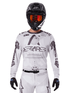 Alpinestars 2025 Racer Hollow Jersey, Motocross-Trikot, Weiß und Schwarz, getragen mit passender weißer MX-Hose und einem schwarzen Helm mit orangefarbener Brille, mit markantem Alpinestars-Branding und Globus-Grafiken, professionelle Offroad-Rennausrüstung