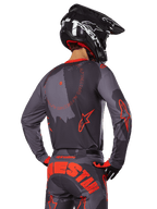 Alpinestars 2025 Racer Hollow Trikot, Rückansicht eines Motocross-Fahrers in Grau Camo und Orange Fluo Ausrüstung, mit passender Camo-Hose mit großem orangefarbenem Alpinestars-Schriftzug und einem schwarzen Helm, professionelles Offroad-Bekleidungsdesign