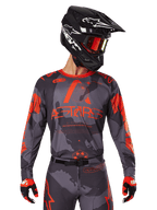 Alpinestars 2025 Racer Hollow Jersey, Motocross-Trikot, Gray Camo und Orange Fluo, getragen mit passendem Ausrüstungsset inklusive Hose und Handschuhen, schwarzer Helm mit weißen Grafiken und Schutzbrille, markantes ASTARS-Branding, schützende Offroad-Rennsportbekleidung