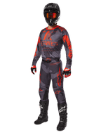 Motocross-Fahrer in kompletter Rennausrüstung, Alpinestars 2025 Racer Hollow Trikot und Hose in Grau Camo Orange Fluo, schwarzer Helm mit orangefarbener Brille, schwarz-rote Handschuhe und schwarze Stiefel mit weißen Logos, abgestimmtes Performance-Outfit für den Offroad-Rennsport