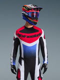 Alpinestars 2026 Techstar Knif Trikot, Motocross-Trikot, Rot, Weiß und Blau, mit markantem Brustlogo und Torso mit Farbverlauf, getragen von einem Fahrer mit passendem mehrfarbigem Helm, blauer Goggle und schwarzen Handschuhen, professionelle Offroad-Rennausrüstung.