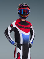 Motocross-Fahrer im Alpinestars 2026 Techstar Knif Trikot in Rot, Weiß und Blau, mit schwarzem Branding auf der Brust, passender Motocross-Hose, abgestimmtem Rennhelm mit blauer Brille und schwarz-weißen Handschuhen, professionelle Offroad-Ausrüstung für den Wettkampf