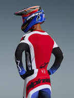 Motocross rider from the back wearing the 2026 Techstar Knif Gear Set, red jersey with white side panels and black sleeves with blue speckled cuffs, red and white pants with black Alpinestars branding across the seat, coordinated blue and red helmet with goggles.