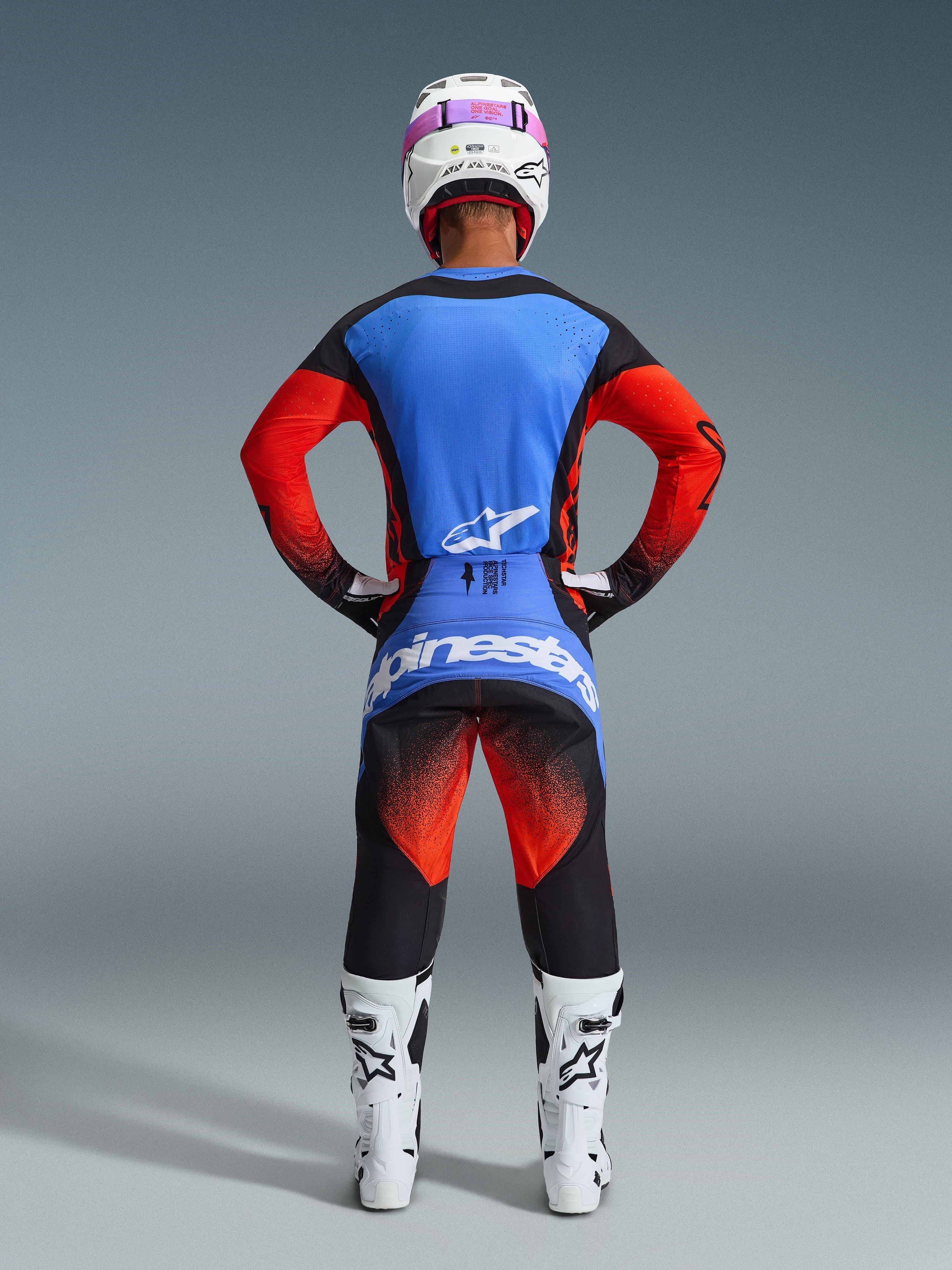Motocross-Fahrer von hinten in kompletter Rennausrüstung, 2026 Techstar Knif Jersey mit blauem Rücken und orangefarbenen Ärmeln, schwarze Hose mit orangefarbenen und blauen Akzenten, weißer Helm mit rosa Brillenband, weiß-schwarze Stiefel, koordiniertes Outfit für Offroad-Rennen